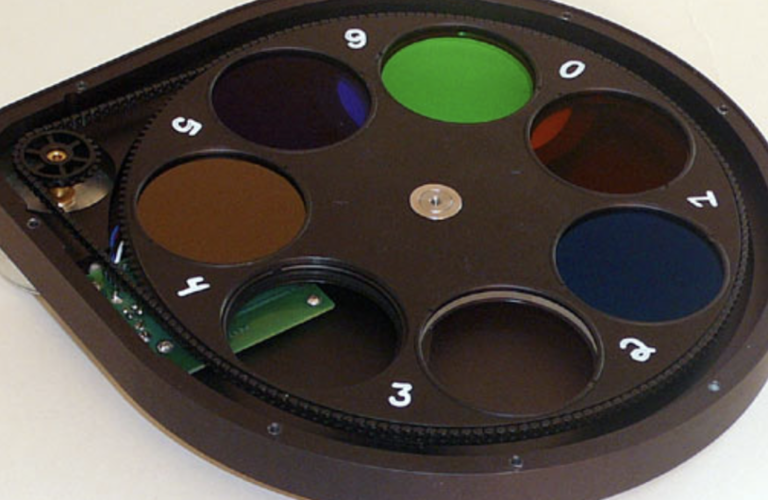 A teardrop-shaped filter wheel is opened up to show the mechanics within. The filter wheel holds a thin rotatable disk that then in turn holds individual circular filters. The filters seen are orange, purple, green, red, and blue. Two holes in this filter wheel are empty.