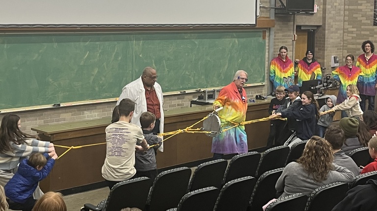  Dale Stille and another demonstrator stand in Van Allen Hall as two groups of children and adults engage in a tug-of-war using yellow ropes. The ropes are attached to a pair of Magdeburg hemispheres—two metal halves held together by a vacuum—to demonstrate the immense strength of atmospheric pressure. Several student assistants in vibrant tie-dye lab coats watch from the side