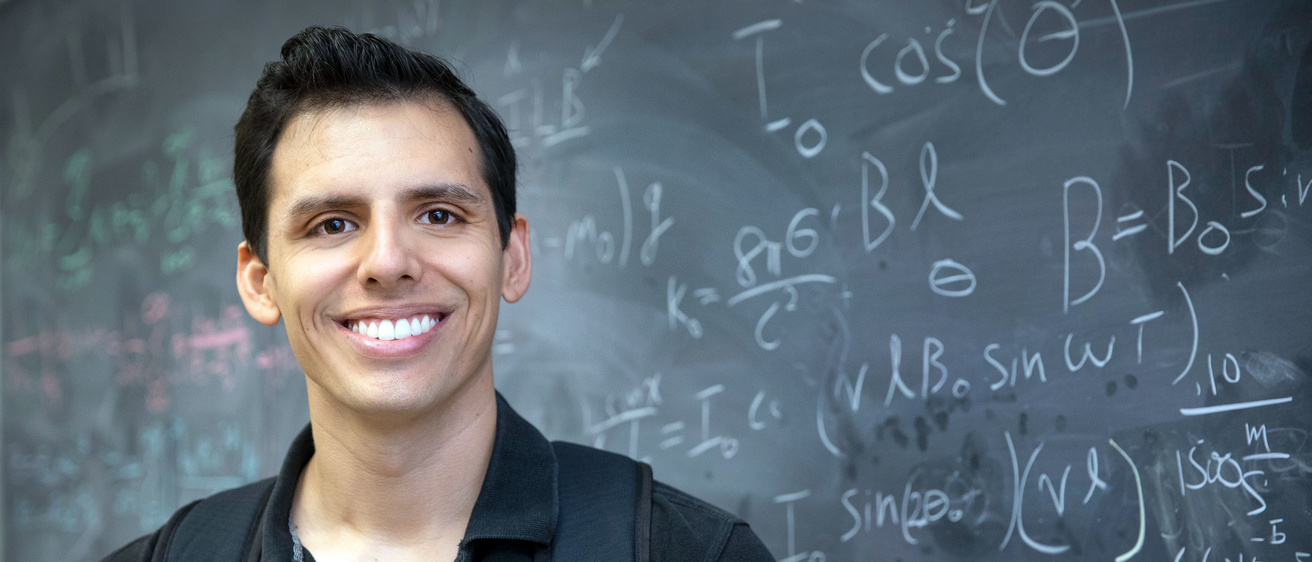 A male Physics student smiling in front of a blackboard with Physics equations written on it.